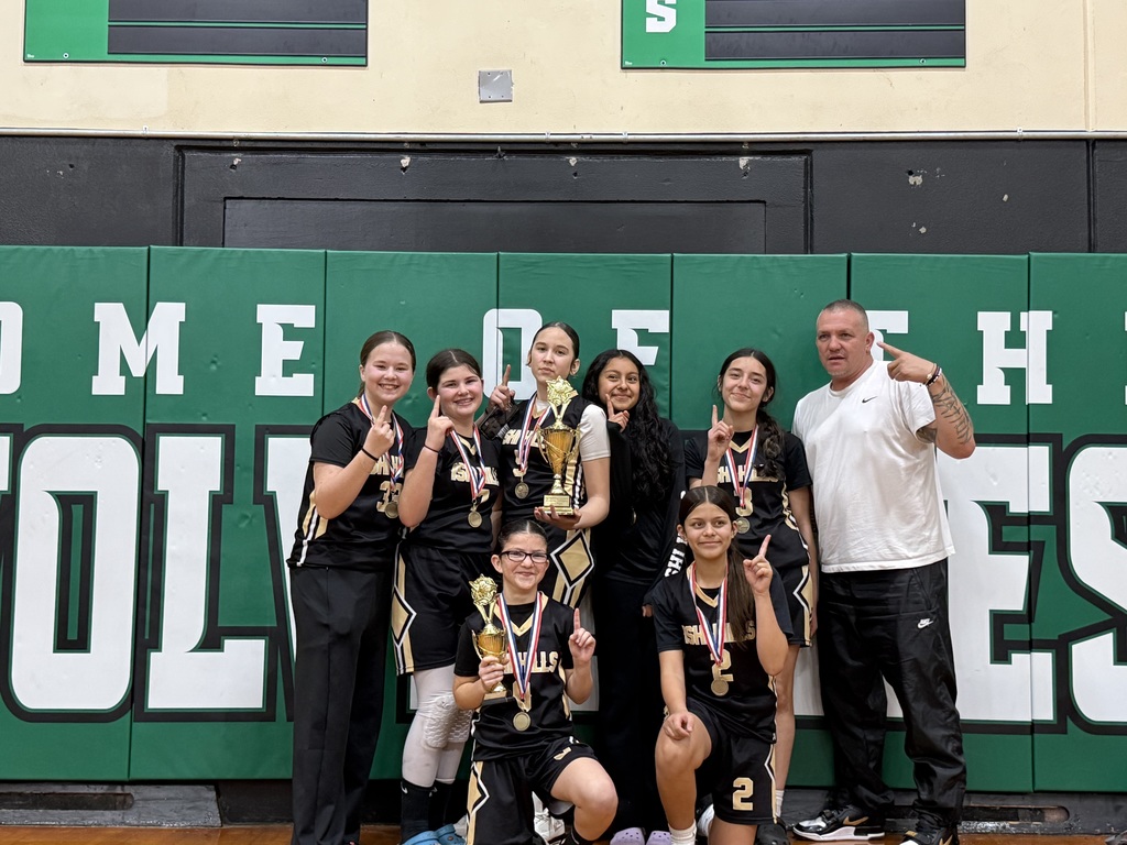 "The Ishi Hills girls basketball team and their coach posing together in front of a 'Home of the Wolves' wall. The players are wearing black and gold uniforms with medals around their necks, holding up two championship trophies and pointing 'number one' signs while celebrating their first-place win."