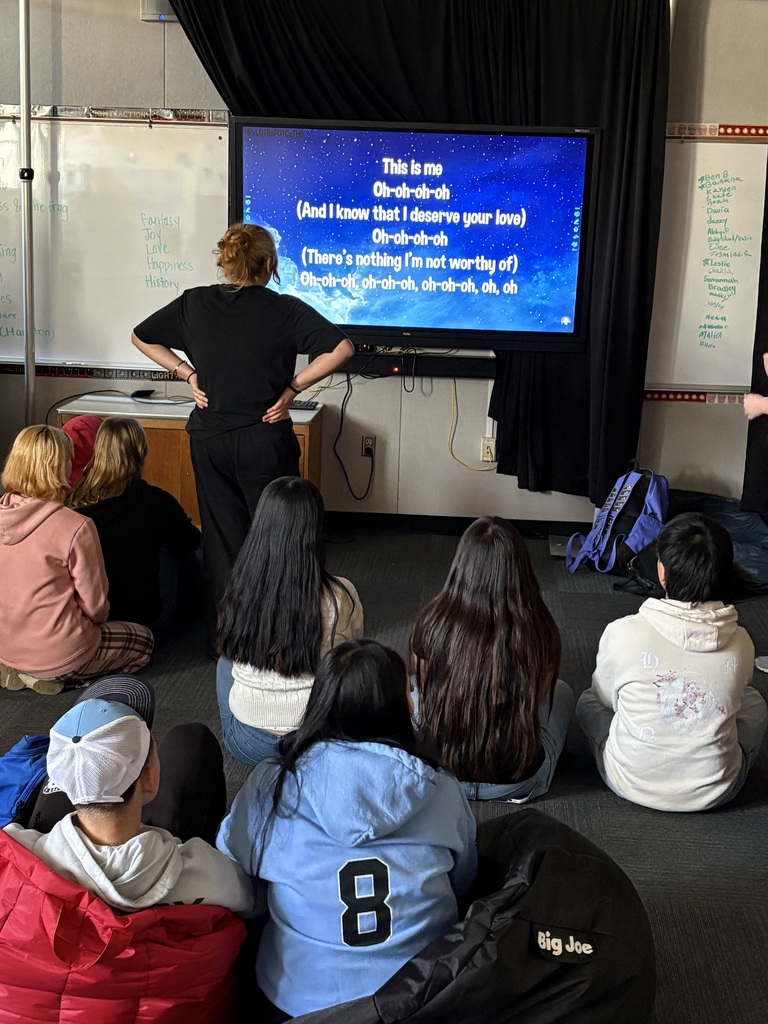 The Focal Point: A large television or monitor displays song lyrics against a starry blue background. The lyrics are from the song "This Is Me" (from The Greatest Showman), featuring the lines: "This is me / Oh-oh-oh-oh / (And I know that I deserve your love)..."  The Participants: * One person, wearing a black t-shirt and black pants, stands at the front with their hands on their hips, looking toward the screen.  A group of about seven students are seated on the carpet in the foreground, viewed from behind. They are wearing casual clothing like hoodies (one light blue hoodie has a large number 8 on the back) and hats.  The Setting: * To the left and right of the screen are whiteboards. The left whiteboard has a list of words including "Fantasy," "Joy," "Love," and "History." The right whiteboard features a list of names.  The room is dimly lit, with dark curtains draped around the screen to create a small "stage" or theater-like atmosphere.