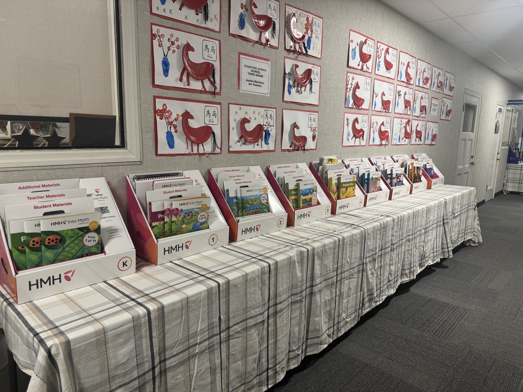 A long, high-angle shot shows a hallway lined with tables covered in a tan and white plaid tablecloth. Arranged in a neat row are approximately ten white HMH Into Math California display kits, each representing a different grade level.  The kits are sequentially numbered in small circles on the bottom right corner, starting with "K" for Kindergarten on the far left, followed by "1," "2," "3," and so on, continuing down the length of the table. Each kit is filled with tiered sections for Additional, Teacher, and Student Materials, featuring colorful workbooks with various illustrations like ladybugs, hot air balloons, and landscapes.