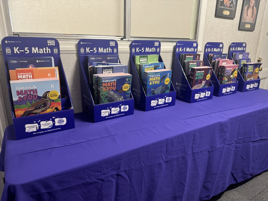 A row of six dark blue California MATH & YOU display stands for elementary education is arranged on a table covered with a solid purple tablecloth. Each stand is labeled "K–5 Math" at the top and contains several spiral-bound textbooks organized by grade level:  Kindergarten (Far Left): Features orange and blue covers with an illustration of a bird (a California quail).  Grade 1: Features blue covers with an illustration of a whale tail under the sea.  Grade 2: Features green and blue covers with an illustration of a breaching whale.  Grade 3: Features dark blue covers with an illustration of a cat.  Grade 4: Features pink and blue covers with an illustration of a bear.  Grade 5 (Far Right): Features dark blue and gold covers with a mountain landscape.