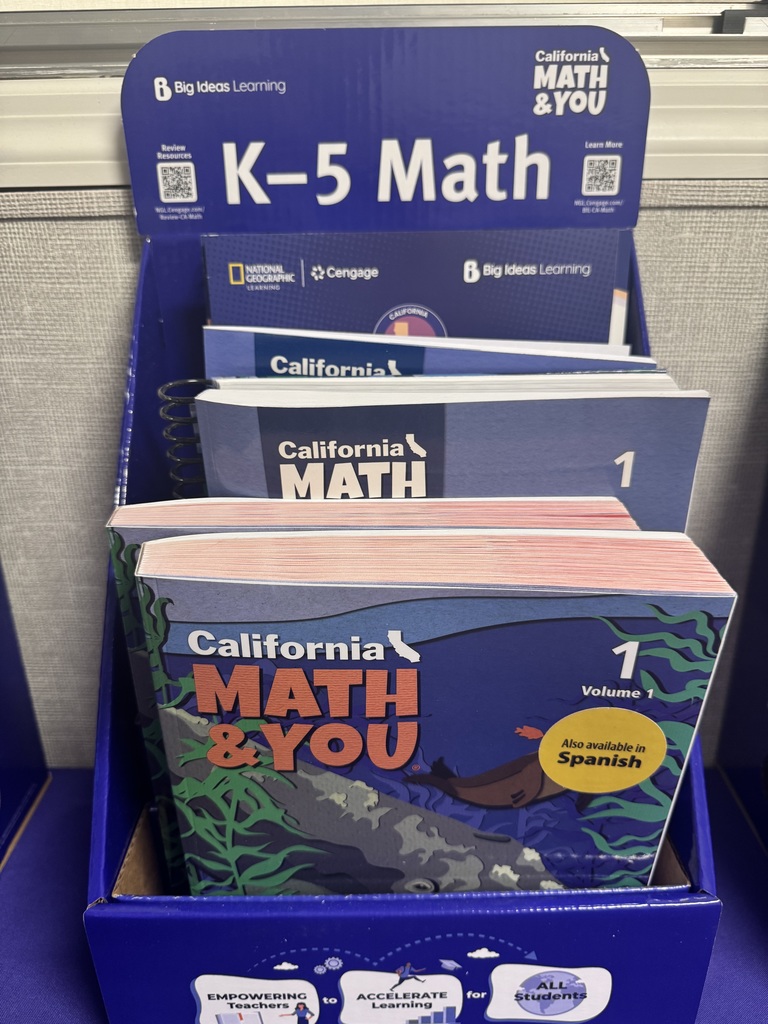 his vertical, close-up shot shows a dark blue California MATH & YOU display stand for Grade 1. The stand is part of the K–5 Math series by Big Ideas Learning, in partnership with National Geographic Learning and Cengage.  The front textbook, Grade 1, Volume 1, features a vibrant underwater scene. The illustration depicts a seal swimming through blue water filled with green kelp and small orange fish. A large yellow circle on the cover indicates that the curriculum is "Also available in Spanish."