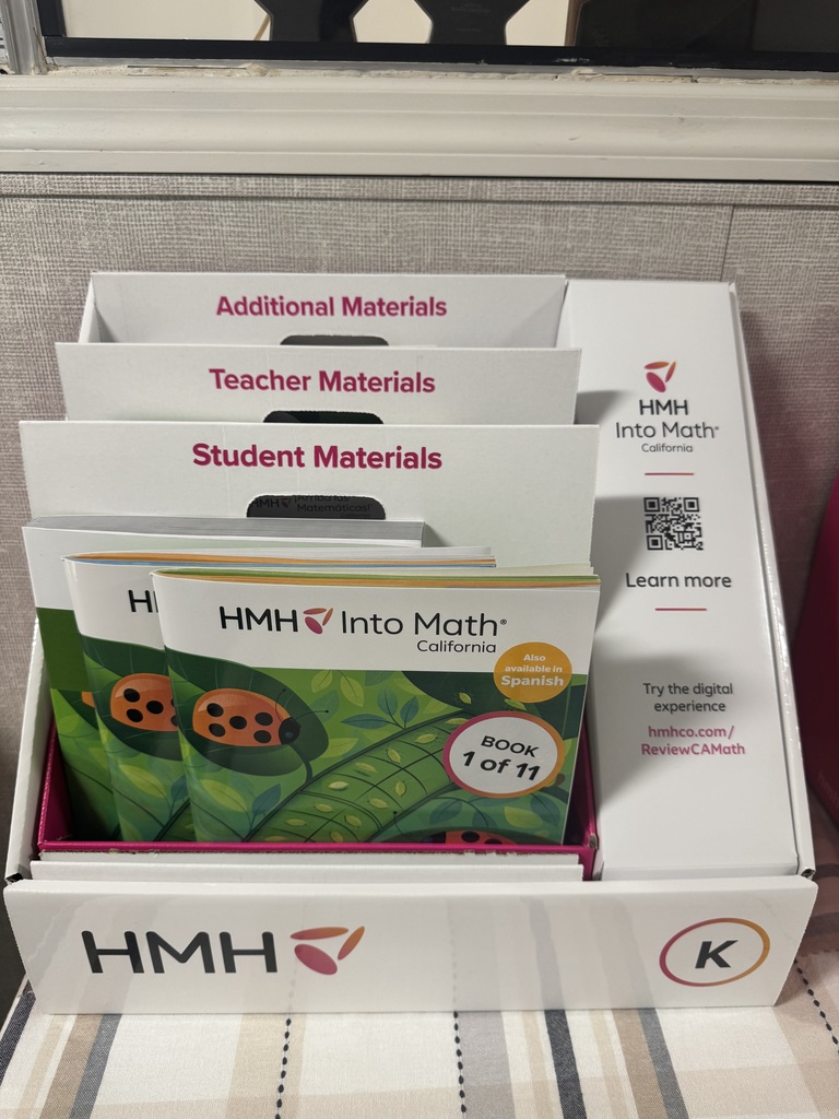 An eye-level, medium shot shows an HMH Into Math California educational display kit sitting on a striped surface. The white cardboard display box is organized into tiered sections labeled in pink text: Additional Materials, Teacher Materials, and Student Materials.