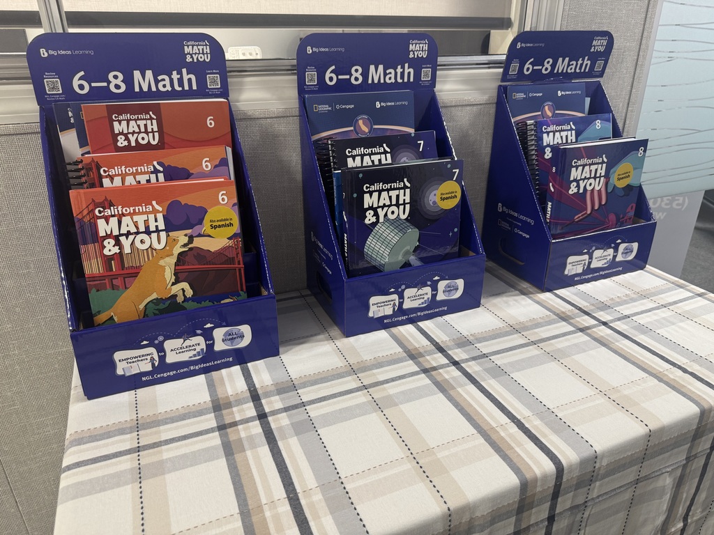 Three dark blue cardboard display stands for the California MATH & YOU curriculum are arranged in a row on a tan, gray, and white plaid tablecloth. Each stand is labeled "6–8 Math" at the top and contains spiral-bound textbooks for a specific grade level:  Grade 6 (Left): Features red and orange covers with an illustration of a dog in front of the Golden Gate Bridge. A yellow circle notes, "Also available in Spanish."  Grade 7 (Middle): Features dark blue covers with an illustration of a satellite. It also includes the Spanish availability notice.  Grade 8 (Right): Features blue and purple covers with an abstract design.