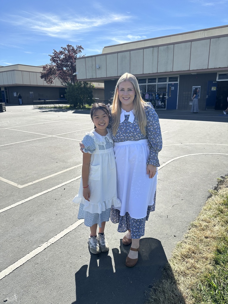 Mrs. Stark and a 5th-grade student at Wyandotte Academy posing together outdoors on a sunny day. Both are dressed in colonial-style outfits, featuring blue floral dresses and white aprons. They are smiling warmly for the camera on the school’s blacktop.