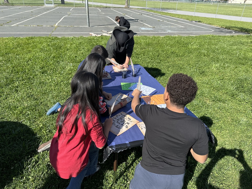 A group of elementary students sitting at a blue outdoor table on a sunny school lawn. They are practicing calligraphy on parchment paper using white feather quills and inkwells as part of a colonial history lesson.