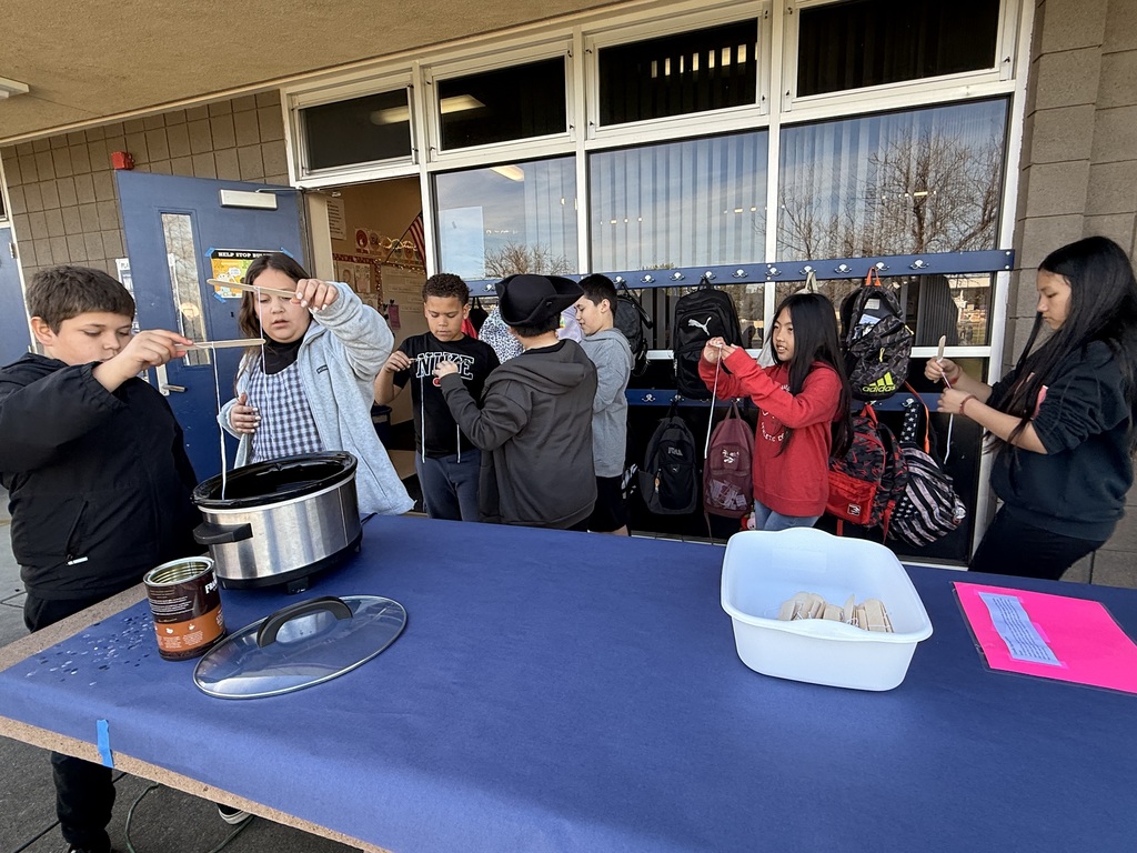 A group of 5th-grade students standing around an outdoor table, practicing the colonial craft of candle dipping. Two students are carefully lowering wicks attached to wooden sticks into a slow cooker filled with wax, while others stand nearby preparing their materials.