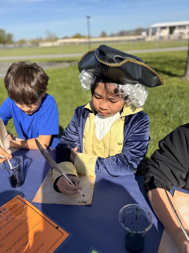 A student at Wyandotte Academy dressed in full colonial attire, including a black tricorn hat with gold trim, a white powdered wig, and a blue and yellow velvet frock coat. He is sitting outdoors at a blue table, focused on writing with a quill and ink on parchment paper.