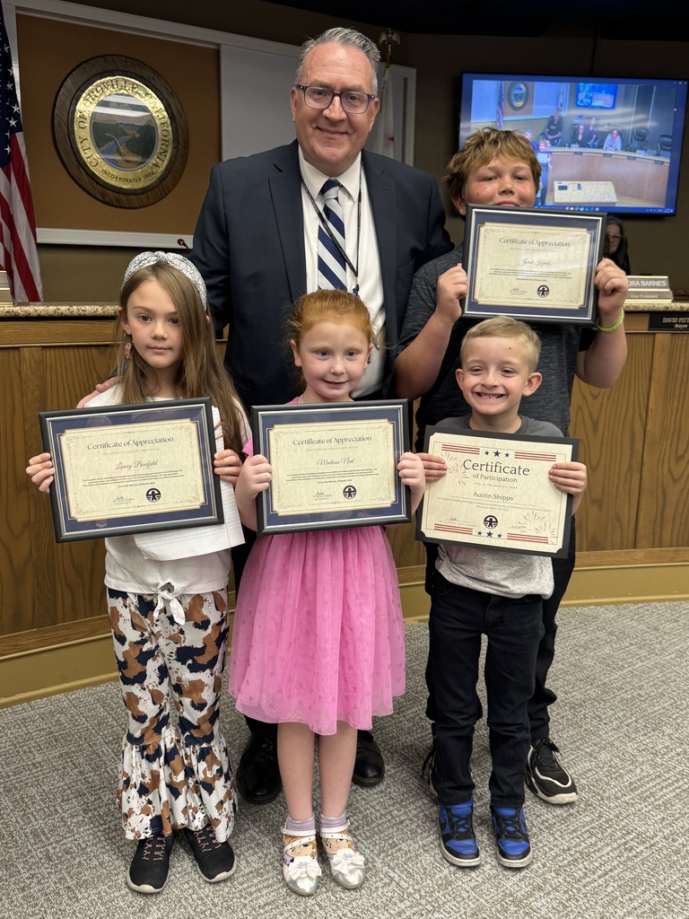 A group photo at a City of Oroville Board meeting featuring Superintendent Holtom  in a suit and tie standing behind four young students who are proudly holding framed certificates. From left to right, the students include a girl in patterned flared pants, a girl in a pink dress, a young boy in a grey shirt, and a taller boy in a black shirt. They are standing in front of a wooden dais with the City of Oroville seal visible on the wall behind them.