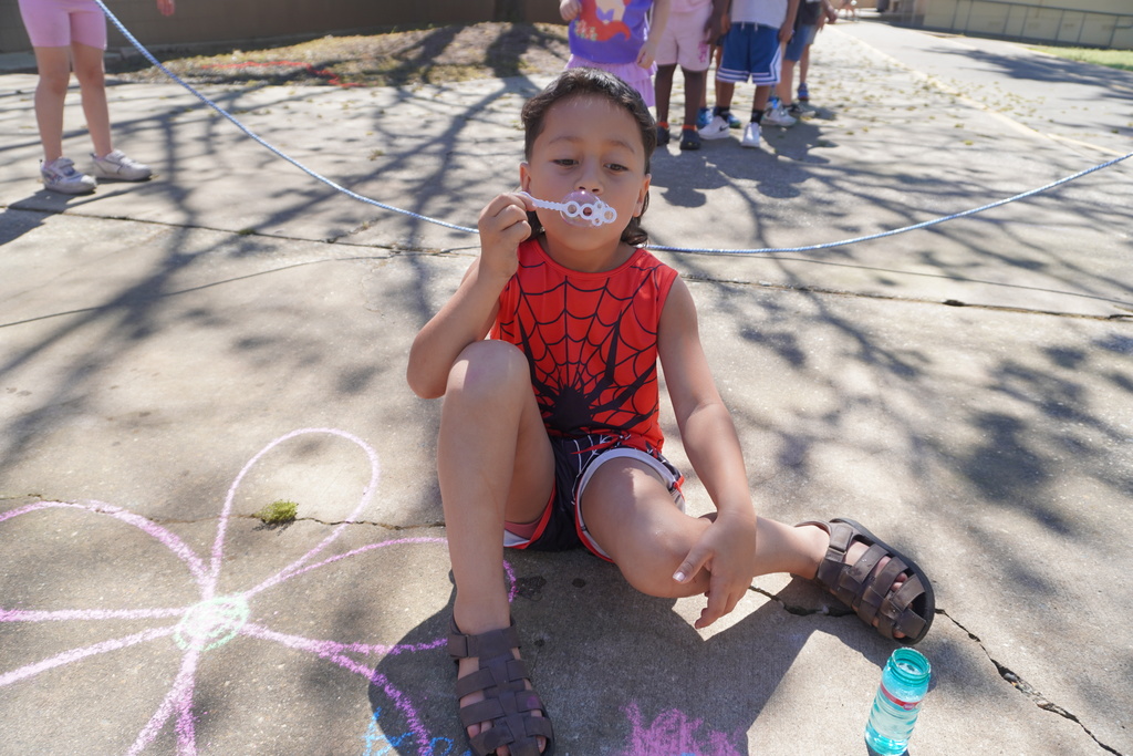 A young boy in an orange Spider-Man tank top and brown sandals sits on a sunny concrete courtyard, blowing bubbles with a white wand. To his left, a large pink flower is drawn on the ground in sidewalk chalk. In the background, other children are lined up behind a blue jump rope, with the dappled shadows of trees falling across the scene.