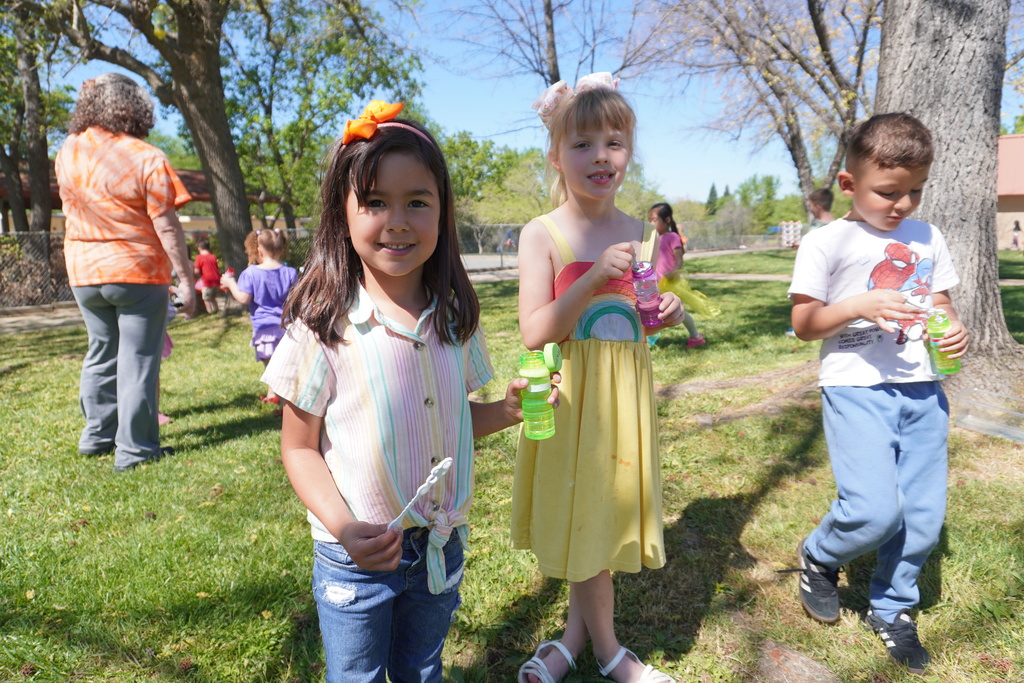 A group of young children are enjoying a sunny day outdoors on a grassy field. In the foreground, two young girls stand side-by-side; one wears a striped shirt and a bright orange hair bow, holding a bubble wand, while the other wears a yellow dress with a rainbow pattern and holds a pink bubble bottle. To their right, a young boy in a Spider-Man t-shirt is focused on his own bottle of bubbles. In the background, other children and an adult in an orange tie-dye shirt are scattered across the lawn under the shade of leafy trees.
