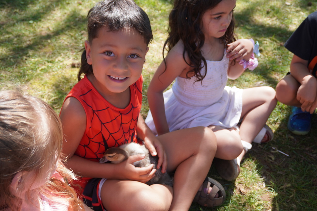 A smiling young boy in an orange Spider-Man tank top sits on a sunny patch of grass, gently holding a small, fuzzy duckling in his lap. To his right, a young girl in a white dress sits cross-legged, looking on, while another child's blonde hair is visible in the lower-left foreground. The group is gathered outdoors under the dappled shade of trees.