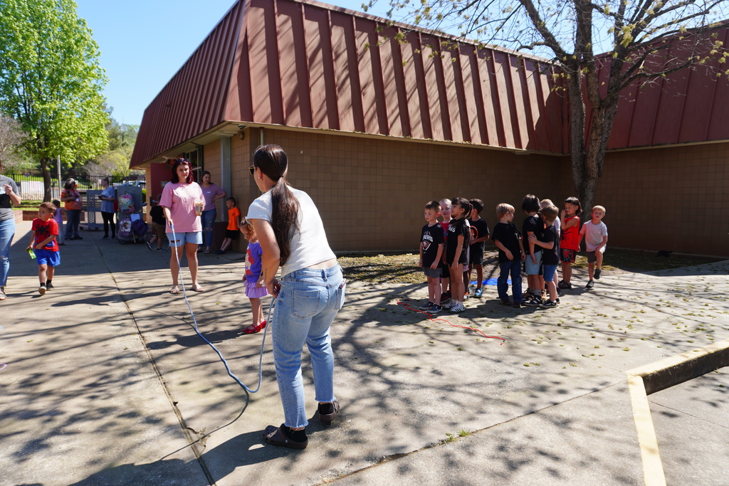 A wide shot of a sunny school courtyard at Ophir Elementary, where two adults are holding the ends of a long blue jump rope for a line of eager kindergarteners. The children are waiting their turn in front of a brown brick building with a distinct red-brown sloped roof. The concrete ground is dappled with shadows from nearby trees, and other students and staff can be seen in the background enjoying the outdoor activities.