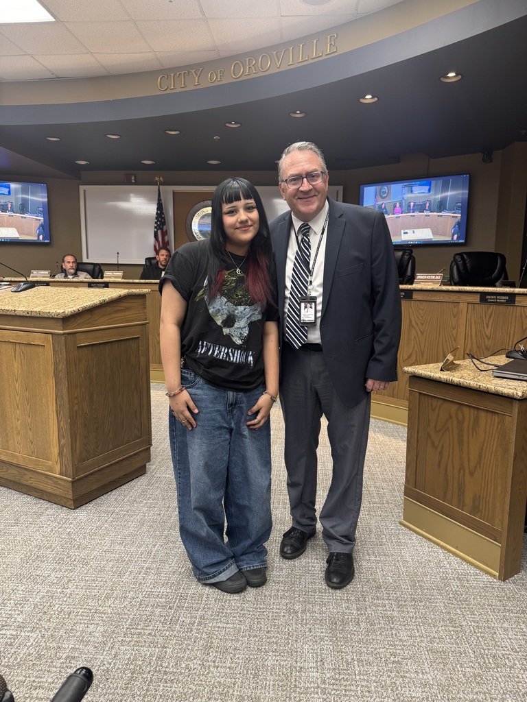 A young woman with dark hair and a black "Aftershock" t-shirt stands next to a smiling man in a navy suit and striped tie inside a government chambers room. They are posing together in front of a curved wooden dais with the words "City of Oroville" visible on the wall behind them. American flags and digital monitors showing a meeting are visible in the background.