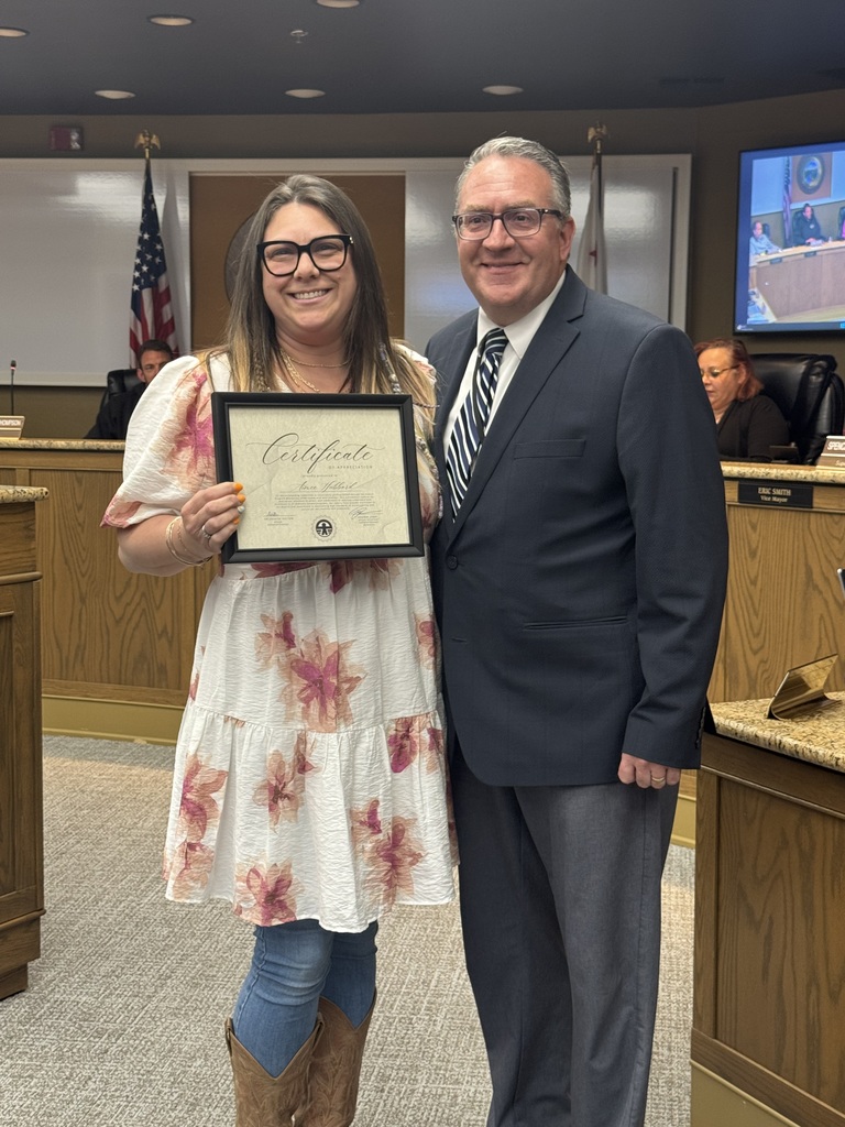 A woman with glasses and a floral print dress stands next to a man in a navy suit and striped tie. She is smiling and holding a framed "Certificate of Appreciation" presented by the school district. They are standing inside the City of Oroville council chambers, with a wooden dais and an American flag visible in the background.