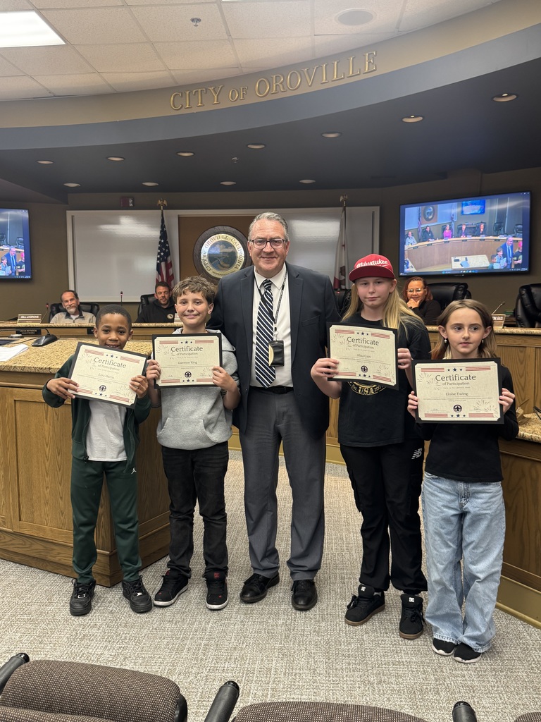 A group photo in the City of Oroville council chambers featuring four elementary students from Oakdale Heights and a man in a suit, likely a school official. Each student is smiling and proudly holding a "Certificate of Participation" for leading the Pledge of Allegiance. The students are dressed casually in hoodies, t-shirts, and jeans, with one wearing a red Milwaukee cap. They are standing in front of a wooden dais with the city seal and American flags visible in the background.