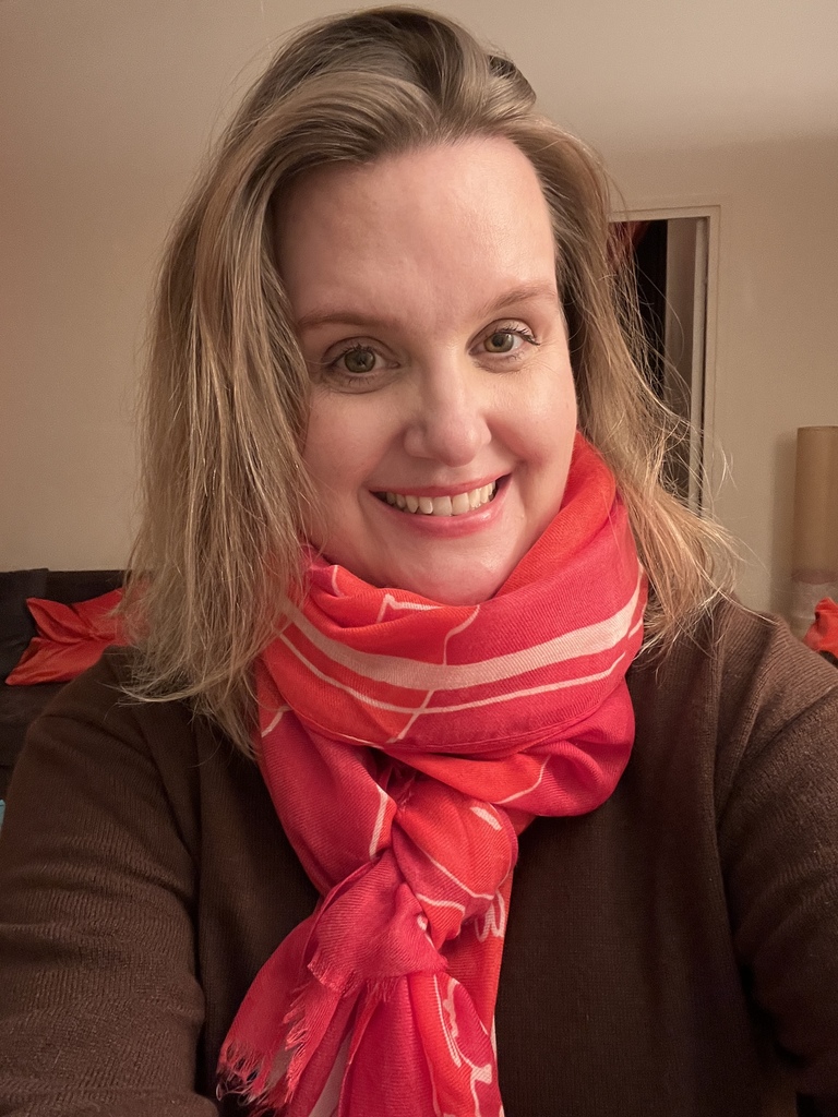 A smiling woman with blonde hair, wearing a brown sweater and a vibrant pink and orange patterned scarf tied around her neck. She is positioned in a brightly lit indoor setting.