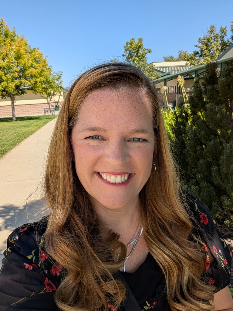 A headshot of a smiling woman with long strawberry-blonde hair and blue eyes, standing outdoors on a sunny day. She is wearing a black blouse with a red floral pattern and a silver necklace. In the background, there is a paved school walkway, green trees, and school buildings under a clear blue sky.
