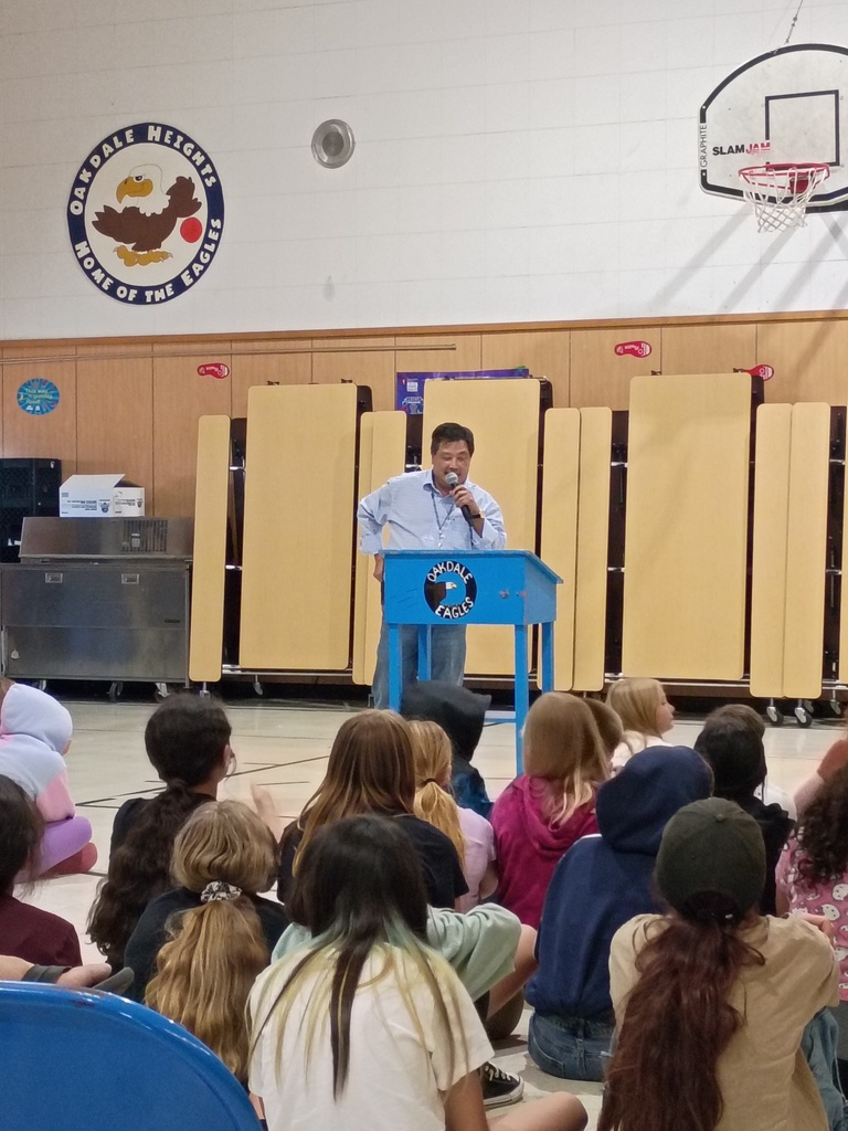 An indoor assembly at Oakdale Heights Elementary School. A man in a light blue button-down shirt stands at a bright blue podium with the "Oakdale Eagles" logo, speaking into a microphone. He is facing a large group of students who are sitting on the gymnasium floor with their backs to the camera. The background features a wooden wall with folded cafeteria tables, a basketball hoop, and a large circular wall decal that reads "Oakdale Heights - Home of the Eagles" featuring a cartoon eagle.