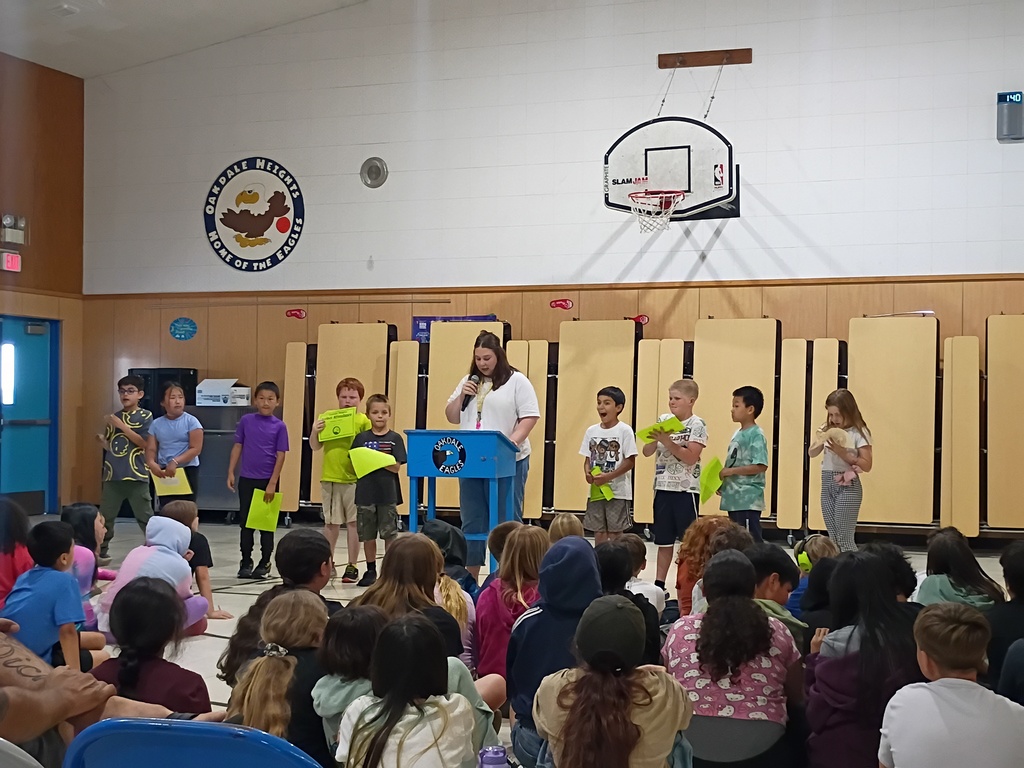 A school official stands at a blue "Oakdale Eagles" podium in a gymnasium, addressing a large group of students. Behind her, a line of nine younger students stand holding bright green and yellow award certificates. The audience is seated on the floor in the foreground, facing the presentation. The gymnasium wall features the Oakdale Heights eagle mascot logo, a basketball hoop, and folded tan cafeteria tables.