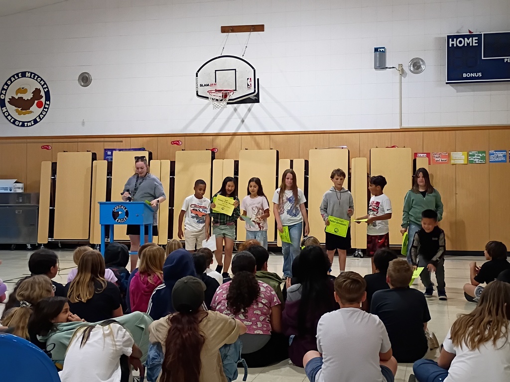 A group of eight elementary students stands in a line at the front of a gymnasium during an awards assembly at Oakdale Heights. Several students are holding bright yellow certificates, including one that clearly reads "Perfect Attendance." To the left, a female staff member in a grey t-shirt stands at a blue "Oakdale Eagles" podium holding a microphone. An audience of students sits on the floor in the foreground, watching the presentation. The background features a basketball hoop, a scoreboard, and the school's eagle mascot logo on the wall.