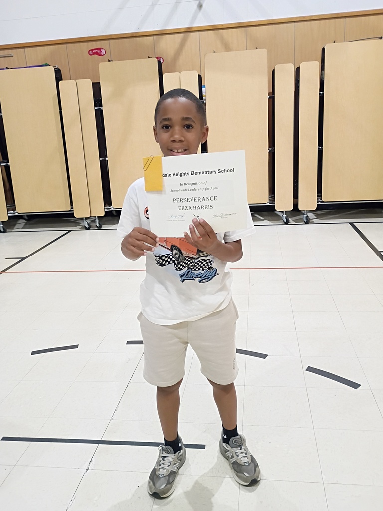 A young boy stands proudly in a school gymnasium, holding up an award certificate from Oakdale Heights Elementary School. The certificate recognizes "ERZA HARRIS" for "School-wide Leadership for April" in the category of "PERSEVERANCE." He is wearing a white graphic t-shirt with a racing car design, tan shorts, and grey sneakers. The background shows the polished gym floor and several folded tan cafeteria tables lined up against the wall.