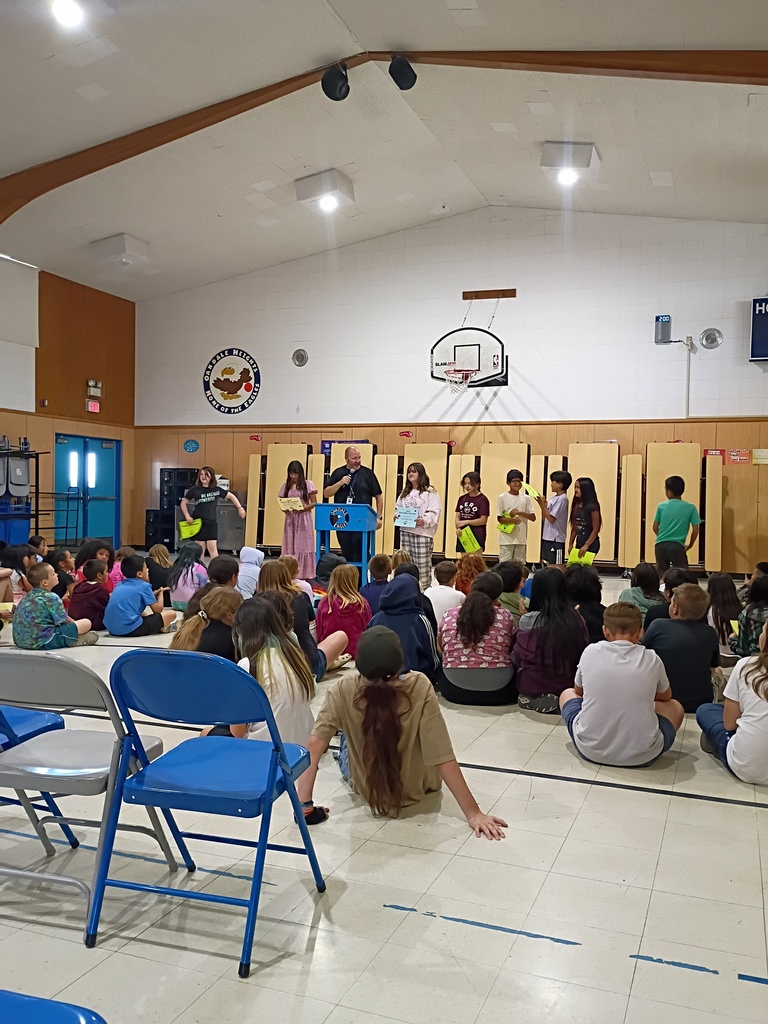 A wide-angle shot of an awards assembly inside the Oakdale Heights gymnasium. A man in a black polo shirt speaks into a microphone at a blue "Oakdale Eagles" podium, standing alongside a group of eight older students who are being recognized. Some students hold yellow and blue certificates. A large audience of students is seated on the floor in the foreground, with blue and grey folding chairs visible in the bottom-left corner. The gym's vaulted ceiling, basketball hoop, and school mascot logo are visible in the background.