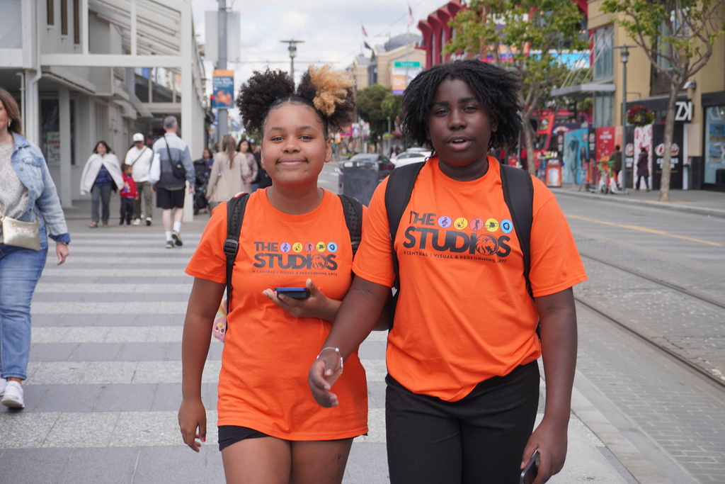 Two middle school students wearing matching bright orange t-shirts that read "THE STUDIOS @ CENTRAL | VISUAL & PERFORMING ARTS" walk side-by-side down a wide pedestrian sidewalk in San Francisco. They are both wearing backpacks, and the student on the left holds a smartphone while smiling at the camera. In the background, other pedestrians walk along the street, which features city buildings, shops, and a set of streetcar tracks running parallel to the sidewalk.