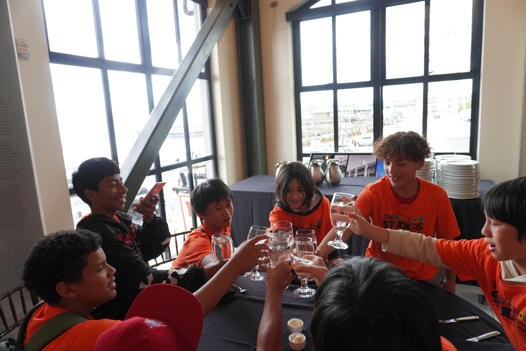 A group of middle school students, several wearing matching orange t-shirts that read "THE STUDIOS," are gathered around a table with a black tablecloth inside a brightly lit restaurant with large windows. The students are smiling and laughing as they clink their water glasses together in a celebratory toast. A stack of white plates and metal pitchers are visible on a buffet table in the background.