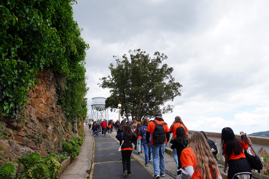 A large group of students and chaperones, many wearing matching bright orange t-shirts, walk up a paved, inclined pathway on Alcatraz Island. The path is lined on the left by a steep rock wall covered in dense green ivy and succulents, and on the right by a low concrete railing. In the distance ahead stands a large tree and the historic Alcatraz water tower under a cloudy, overcast sky.