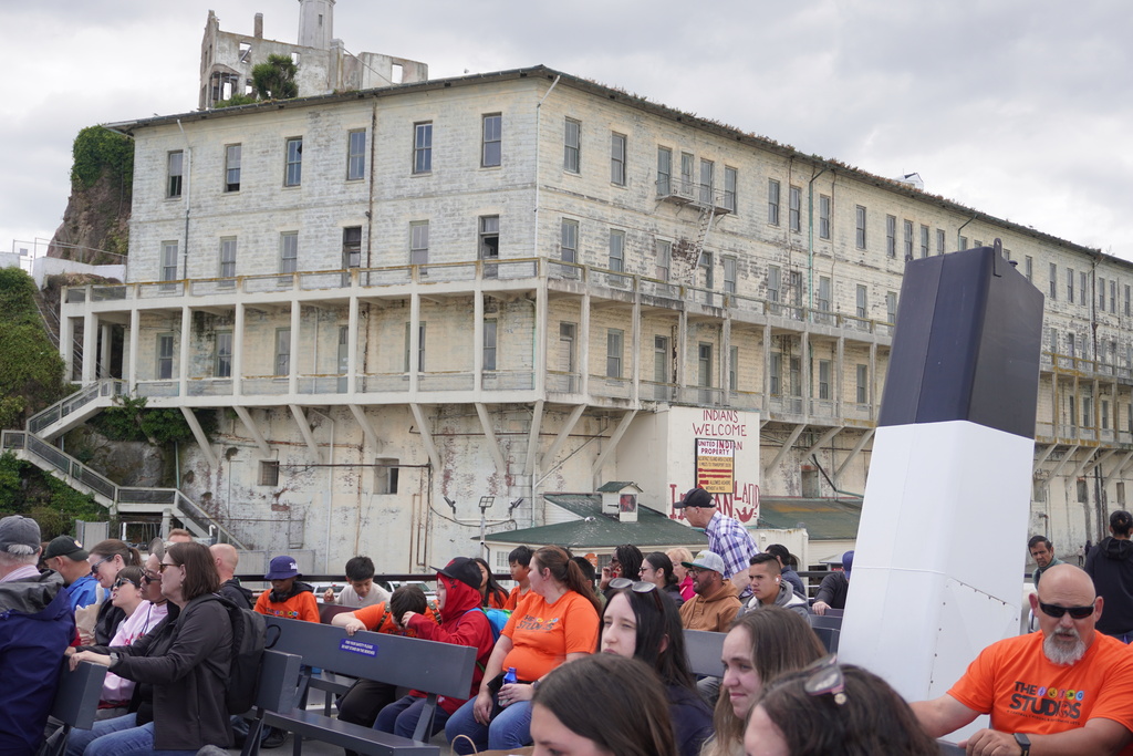 A large group of students and adult chaperones sit on outdoor benches aboard a ferry boat, with many wearing matching bright orange "THE STUDIOS" t-shirts. In the background, the massive, weathered, multi-story historic barracks building on Alcatraz Island towers over the dock. The building features peeling cream paint, rows of windows, and the famous historic "INDIANS WELCOME" graffiti painted on the lower wall beneath a cloudy sky.