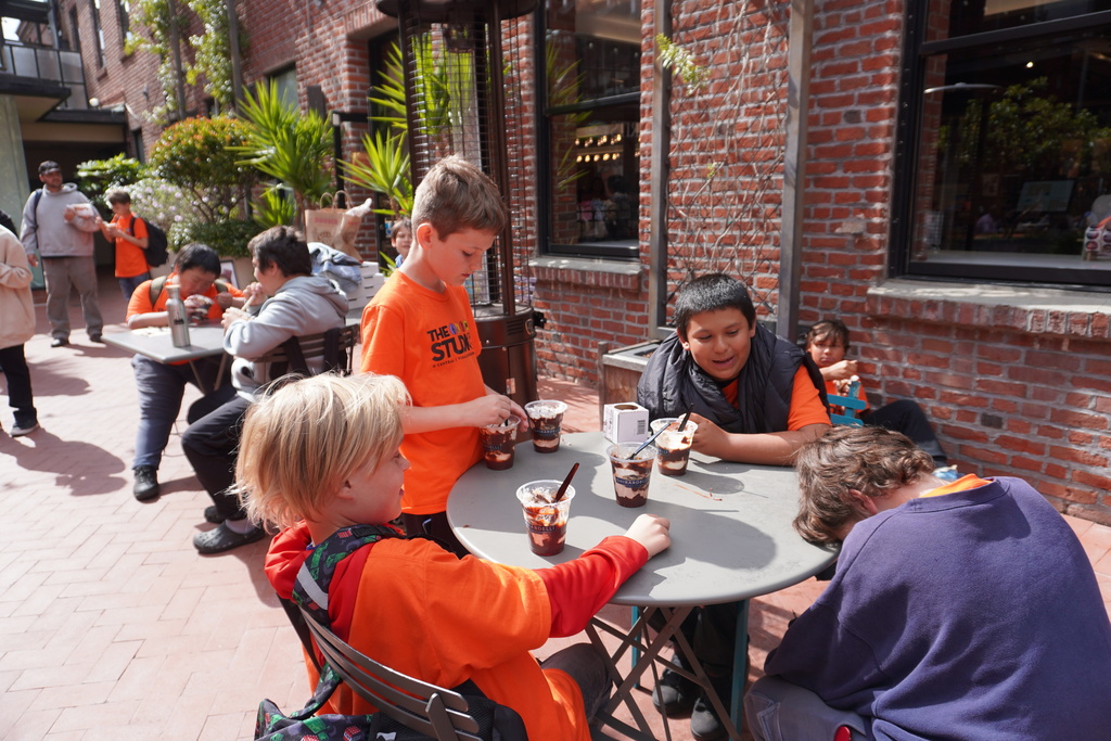 Several middle school students are sitting and standing around a small grey patio table in an outdoor brick courtyard, enjoying ice cream sundaes in clear plastic cups from Ghirardelli. A few of the students wear bright orange "THE STUDIOS" t-shirts. In the background, other students are seated at tables or walking through the courtyard, which is lined with brick buildings, windows, and potted green plants under bright sunlight.