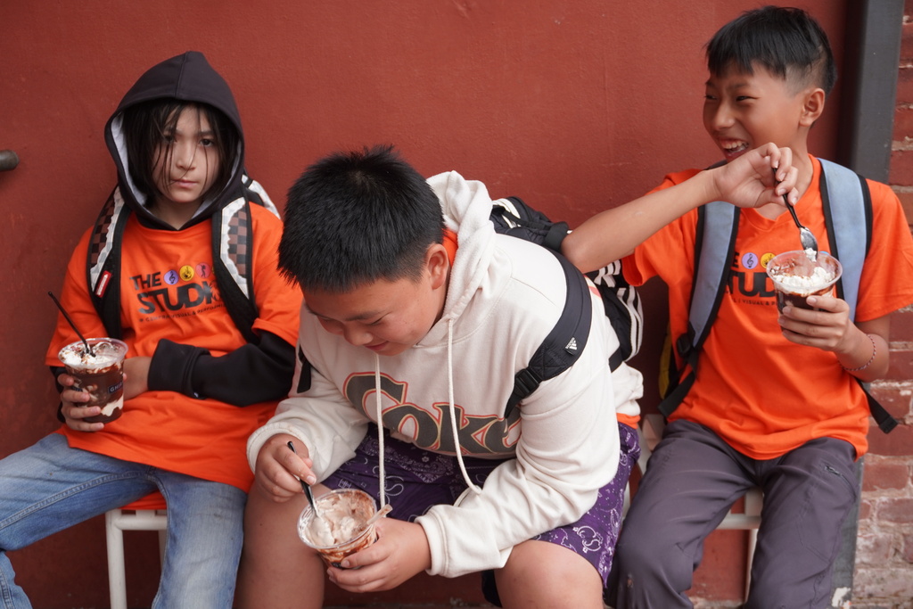 Three middle school students sit together against a reddish-brown wall, smiling and laughing as they eat ice cream sundaes from clear plastic cups. Two of the students wear matching bright orange t-shirts with "THE STUDIOS" logo, while the student in the center wears a white hoodie and purple patterned shorts. They are holding plastic spoons and enjoying their treats, likely at Ghirardelli Square.