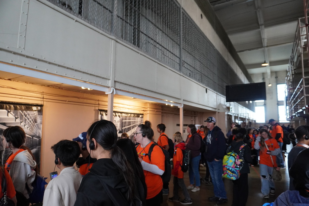 A large group of middle school students and adult chaperones stand inside a wide corridor of a historic prison, likely Alcatraz. Many of the students are wearing bright orange t-shirts and have audio tour headphones on. They are looking at exhibits and historical photographs mounted along the wall under a low ceiling mezzanine. The upper level features high concrete walls and barred window areas, with a long, open hallway stretching into the background under overhead lighting.