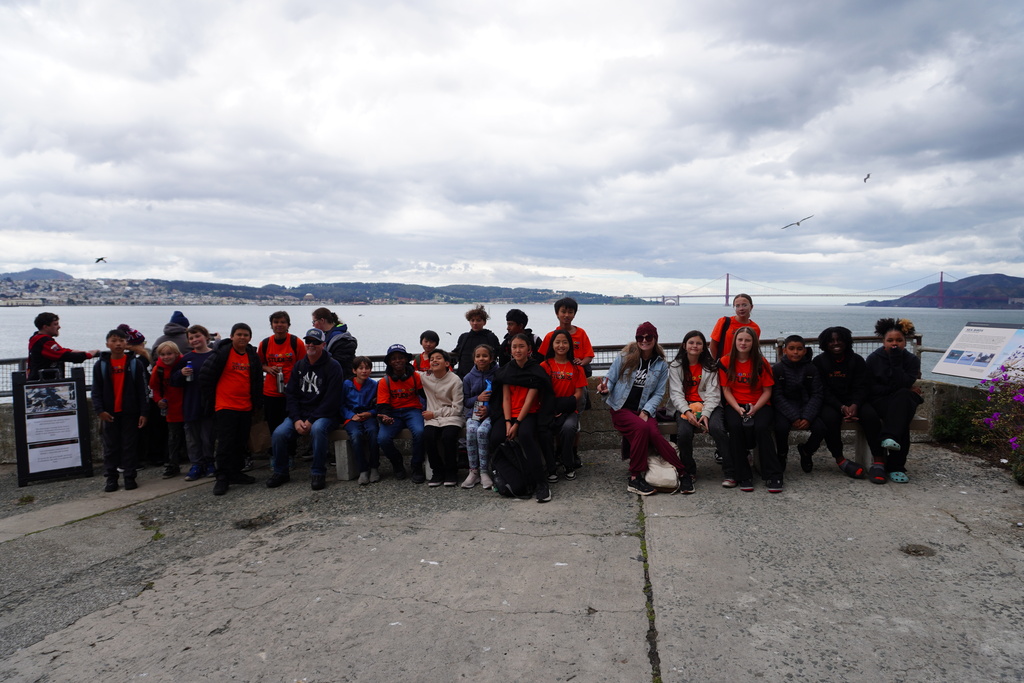 A large group of middle school students and adult chaperones pose together for a photo on an outdoor concrete terrace at Alcatraz Island. Many of the students are wearing matching bright orange t-shirts that say "THE STUDIOS." They are lined up across a low concrete wall and bench, with some sitting and others standing. In the background, the wide, grey-blue water of the bay stretches toward the horizon under a cloudy sky, with the Golden Gate Bridge faintly visible in the distance to the right.