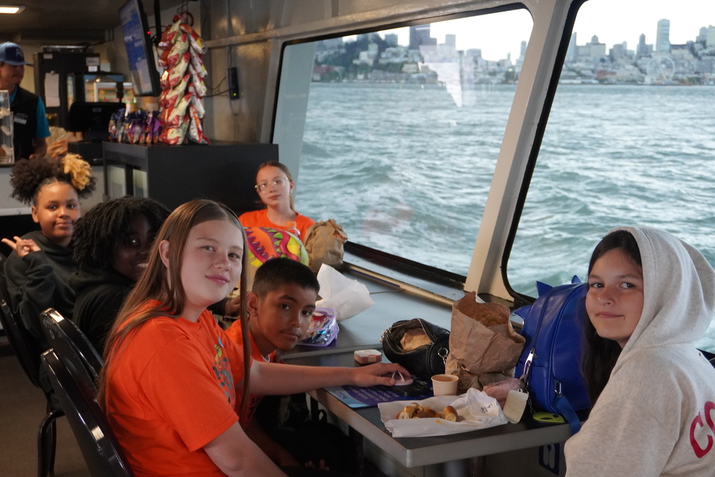 A group of middle school students, some wearing bright orange t-shirts, are sitting at tables inside a ferry boat. Large windows next to them look out over the choppy gray-blue water of a bay toward a distant city skyline under an overcast sky. Some food and bags sit on the tables, and a snack bar counter is visible in the background on the left.