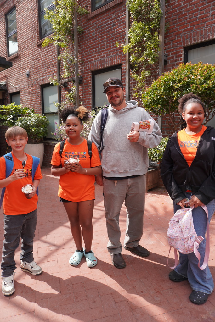 An adult male chaperone stands smiling in the center of three middle school students in an outdoor brick courtyard at Ghirardelli Square. The students are wearing matching bright orange t-shirts that read "THE STUDIOS," with the boy on the left holding a plastic cup of ice cream and a spoon. The chaperone wears a grey hoodie, sunglasses on his hat, and holds a white paper bag from Boudin Bakery. A classic brick building with green climbing vines and large windows serves as the background under bright daylight.