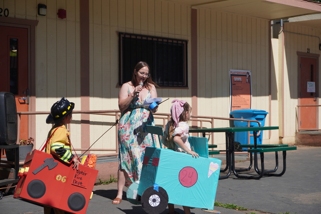 A woman in a floral sundress stands outside a school building speaking into a microphone and holding a paper document. On her left, a young boy dressed in a yellow firefighter costume and black helmet wears a red cardboard fire truck decorated with an axe design and labeled "Ophir Fire Dept...". On her right, a young girl with a pink headscarf stands inside a light blue cardboard truck bearing the number 24 inside a pink circle and a hand-drawn heart. In the background, a beige school building with brown doors, a playground rules sign, green picnic tables, and a blue recycling bin are visible under the bright sunlight.