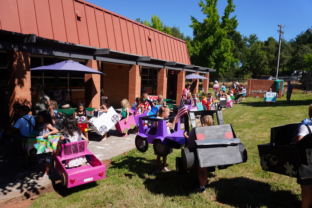A wide shot of a long line of kindergarten students lined up outdoors on a sunny day for a school parade. The children are wearing a wide variety of colorful, homemade cardboard box vehicles. In the foreground on the grass, a girl stands in a large, dark gray or black cardboard truck, while next to her a girl holds a small American flag from inside a bright purple cardboard vehicle. To the left, other children are visible in pink, white, and spray-painted cars near a brick school building with a red metal roof and blue patio umbrellas. The line of kids extends far into the background across the green grass under a clear blue sky.