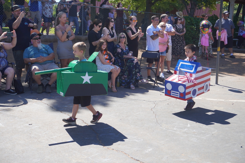 Two young boys participate in an outdoor school parade on a paved playground, walking past a large group of seated and standing spectators. The boy on the left wears a large, green cardboard box military tank complete with a long protruding barrel, a large white star on the side, and a black base mimicking tracks. The boy on the right wears a patriotic cardboard car painted with red and white stripes, blue trim, a small American flag attached to the hood, and a colorful balloon trailing behind it. In the background, family members and onlookers watch the parade from folding chairs and behind a chain-link fence under the bright sun.
