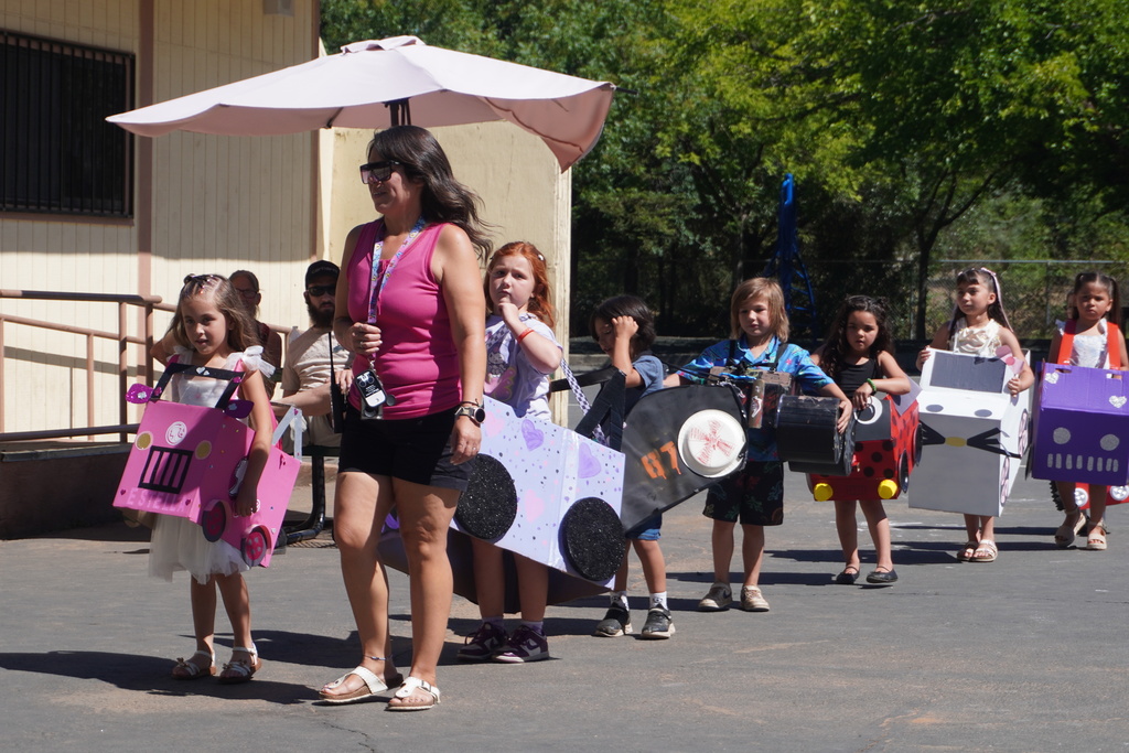 A line of young children walking down an asphalt path during a school parade, led by a woman in a pink tank top and sunglasses holding a light pink umbrella for shade. The children are wearing various creative, homemade cardboard box vehicles. On the left, a girl in a white dress wears a bright pink car labeled "Estella." Behind her, another girl wears a purple car decorated with hearts and glittery black wheels. Further down the line, a boy walks with a black race car bearing the number 87, followed by a girl in a red ladybug-styled car, a girl in a white Hello Kitty-styled car, and a girl in a purple car. Trees and school buildings are visible under the bright sunshine in the background.