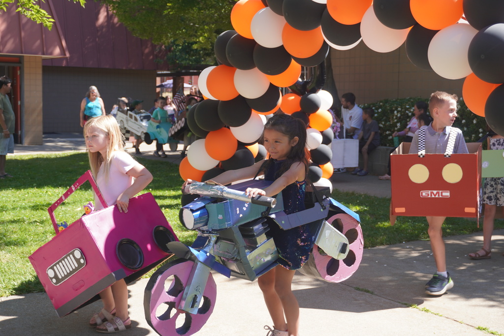 A sunny outdoor school parade featuring young children wearing elaborate homemade vehicles made out of cardboard. In the center, a smiling girl in a sequined dress maneuvers a detailed, three-dimensional cardboard motorcycle painted metallic blue and pink. To her left, a blonde girl walks in a bright pink cardboard Jeep with a hand-drawn grille. To the right, a boy walks inside a red cardboard GMC truck with yellow paper headlights and checkered straps. They are walking along a concrete path on a grassy campus, passing beneath a massive archway of orange, black, and white balloons while onlookers watch in the background.