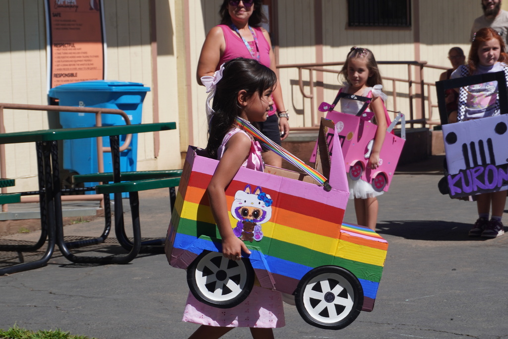 A close-up shot of young girls walking outdoors in a school parade with their custom cardboard box vehicles. In the foreground, a young girl in a pink dress walks forward wearing a car painted with bright, horizontal rainbow stripes and decorated with a cartoon character wearing a Hello Kitty hoodie. Her car has black wheels with white rims and rainbow-colored shoulder straps. Behind her, another girl follows in a bright pink cardboard car, while a third child stands to the right in a purple car featuring a drawn-on grille and the name "Kuron." In the background, a woman in a pink tank top, a blue recycling bin, and a green picnic table are visible next to a school building under the bright sun.