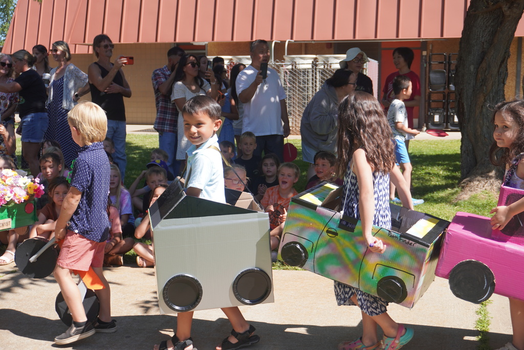 A line of young children participating in a school parade outside on a sunny day. They are wearing custom homemade cars made out of cardboard boxes, complete with paper plate wheels, held up by shoulder straps. In the foreground, a boy smiles at the camera in a light-colored car, followed by a girl wearing a vibrant, tie-dye spray-painted car labeled "Path Finder," and another girl in a bright pink car. In the background, a crowd of parents and onlookers stand on the grass, smiling and taking photos with their phones under a school building with a red awning.