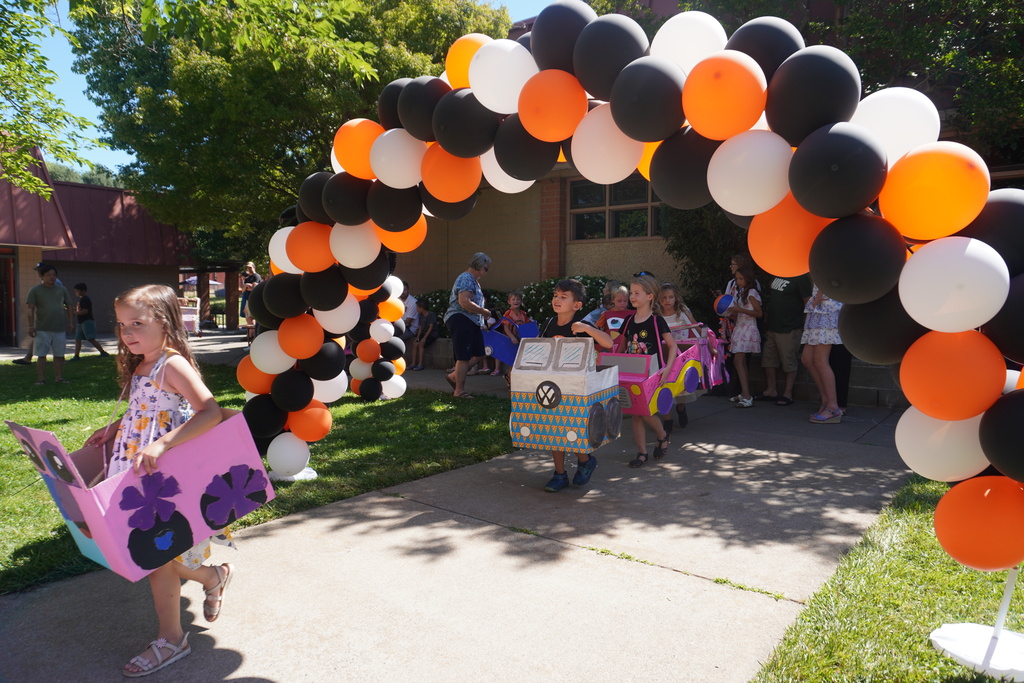 A line of kindergarten students parading down a concrete walkway outdoors beneath a large, overarching balloon arch made of orange, black, and white balloons. The children are wearing homemade cardboard box cars with shoulder straps. In the foreground on the left, a young girl walks forward wearing a bright pink car decorated with large purple paper flowers. Following behind her under the archway is a boy in a cardboard car decorated like a Volkswagen bus with a hand-drawn VW logo, followed by a girl in another pink car. In the background, teachers and family members stand on the grass and pavement watching the parade.