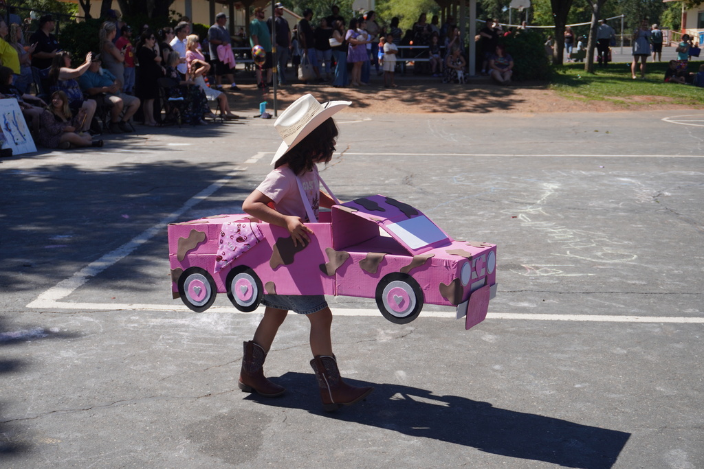 A young girl walks across a paved outdoor playground during a school parade, dressed in a Western theme. She wears a white cowboy hat, a pink t-shirt, a denim skirt, and brown cowboy boots. Suspended by shoulder straps, she wears a large pink cardboard box car decorated with brown cow-print spots, three wheels on the side with small hearts in the center, and a pink bandana draped over the back. In the background, a large crowd of seated and standing parents and onlookers watch from the shaded edges of the playground.