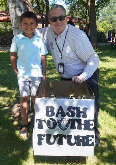 A smiling young boy in a light blue polo shirt and shorts stands next to a man wearing sunglasses and a striped button-down shirt. They are posing outdoors on a sunny, grassy area. In front of them is a large cardboard box decorated with bold, stylized black text that reads "BASH TO THE FUTURE," featuring a Las Vegas Raiders football team logo in the center.