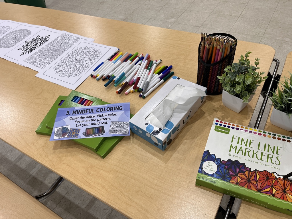 A school table set up as a coloring station. A sign reads "3. Mindful Coloring: Quiet the noise. Pick a color. Focus on the pattern. Let your mind rest." On the table are several printed sheets of intricate mandala coloring designs, a scattered pile of colorful markers, a box of Crayola fine line markers, a mesh cup holding colored pencils, a box of tissues, and two small potted green plants.