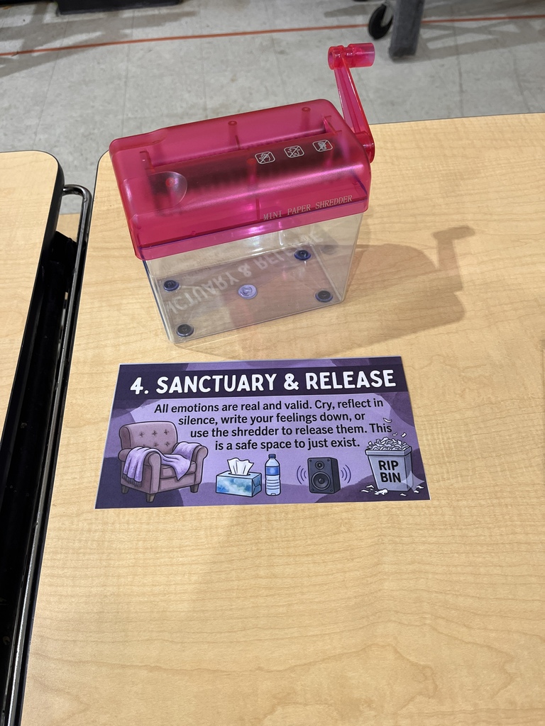 A wooden table featuring a release station. A sign reads "4. Sanctuary & Release: All emotions are real and valid. Cry, reflect in silence, write your feelings down, or use the shredder to release them. This is a safe space to just exist." Sitting directly on top of the sign is a small, clear plastic hand-crank paper shredder with a bright pink lid.