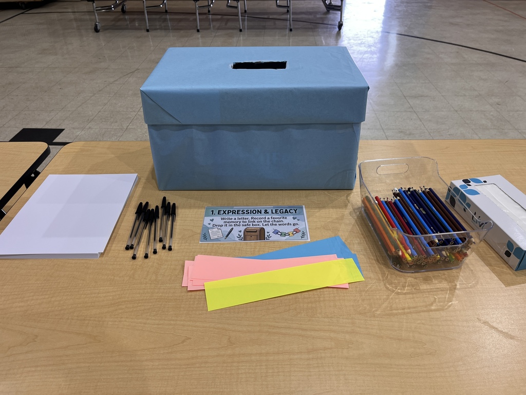 A wooden table set up as a school activity station. A sign in the center reads "1. Expression & Legacy: Write a letter. Record a favorite memory to link on the chain. Drop it in the safe box. Let the words go." Behind the sign is a large light-blue cardboard box with a slot cut into the top. To the left is a stack of white paper and a row of black pens. To the right are strips of blue, pink, and yellow construction paper, a clear plastic container filled with colored pencils, and a box of tissues.