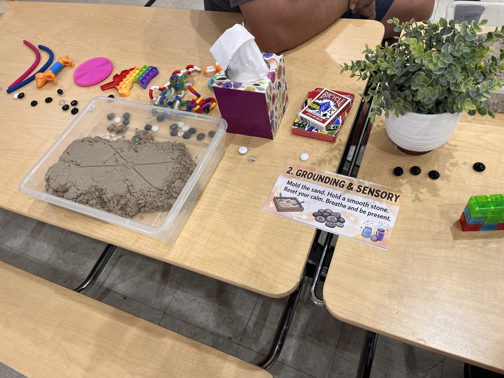 A wooden table set up as a sensory activity station. A sign reads "2. Grounding & Sensory: Mold the sand. Hold a smooth stone. Reset your calm. Breathe and be present." On the table is a clear plastic bin filled with kinetic sand, various colorful pop-it and plastic fidget toys, a deck of cards, small smooth black and white stones, a box of tissues, building blocks, and a small potted green plant.